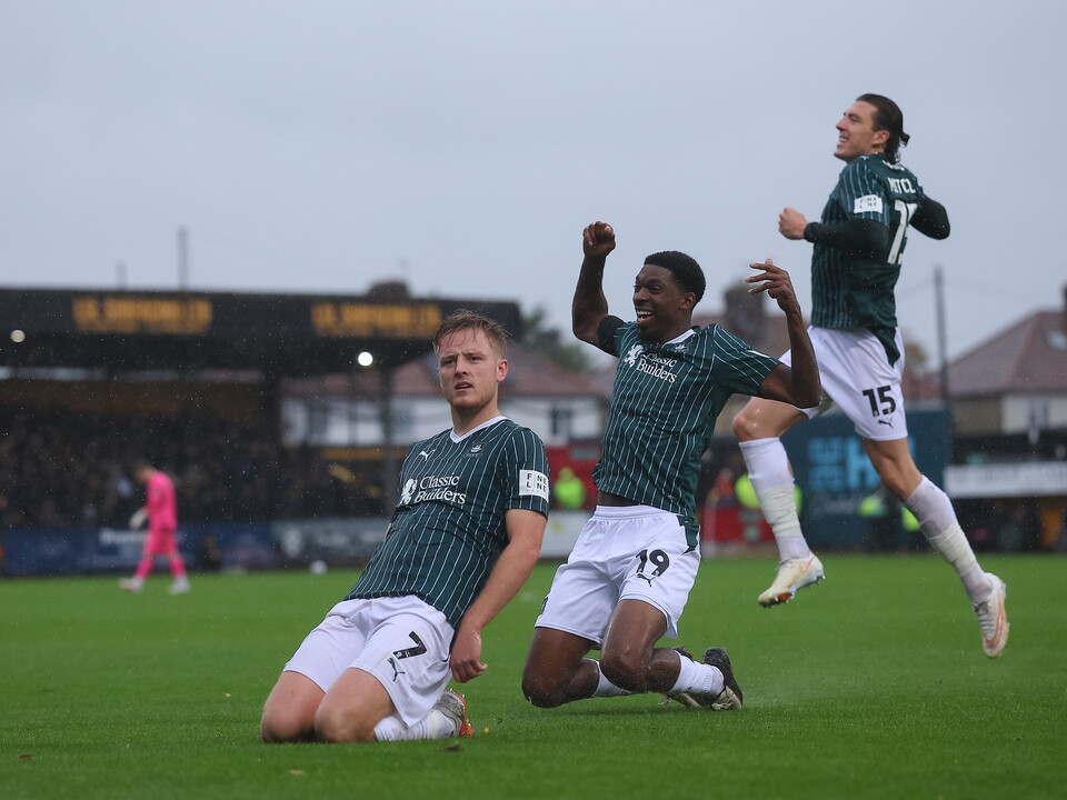 Three footballers, one lying down and two jumping, on a football pitch.