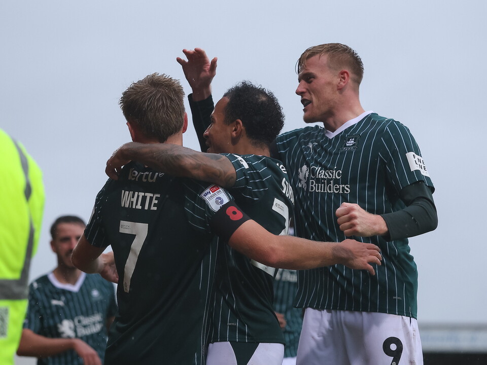 Football players celebrate on the pitch. Player 7 is hugged by two others, one wearing a green and white striped shirt with the number 9.