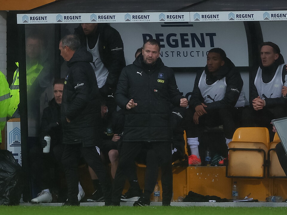 Men in black jackets stand on the sidelines of a football pitch, a coach looks animated.