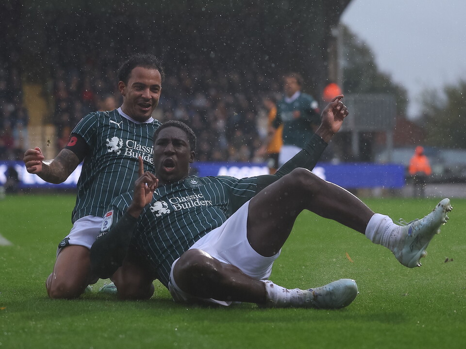 Two players in green and white uniforms fall to the ground on a football pitch.