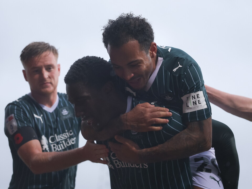 Three football players celebrating, one wearing a green shirt with "NE" and number 22, another with "Classic" on his shirt.