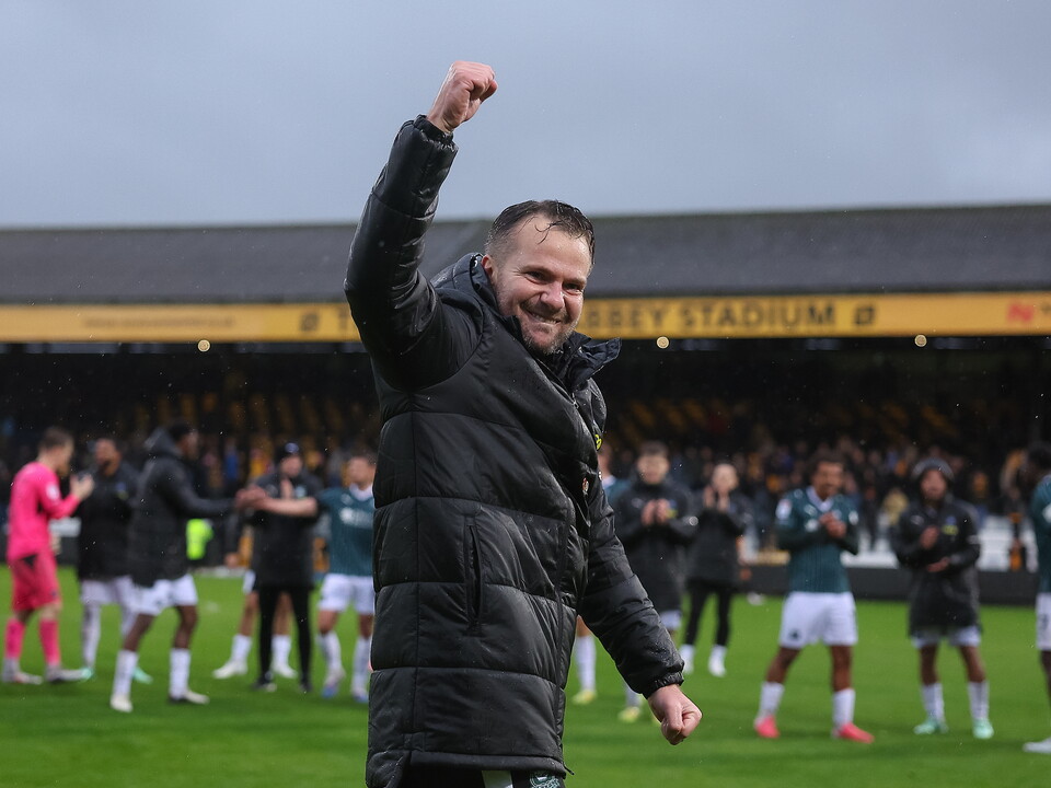 A man wearing a black puffer jacket celebrating in front of a football pitch with players behind