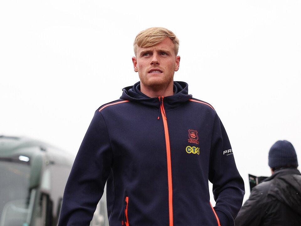A man with blonde hair wearing a navy blue hooded jacket with orange stripes and a logo on the left chest area