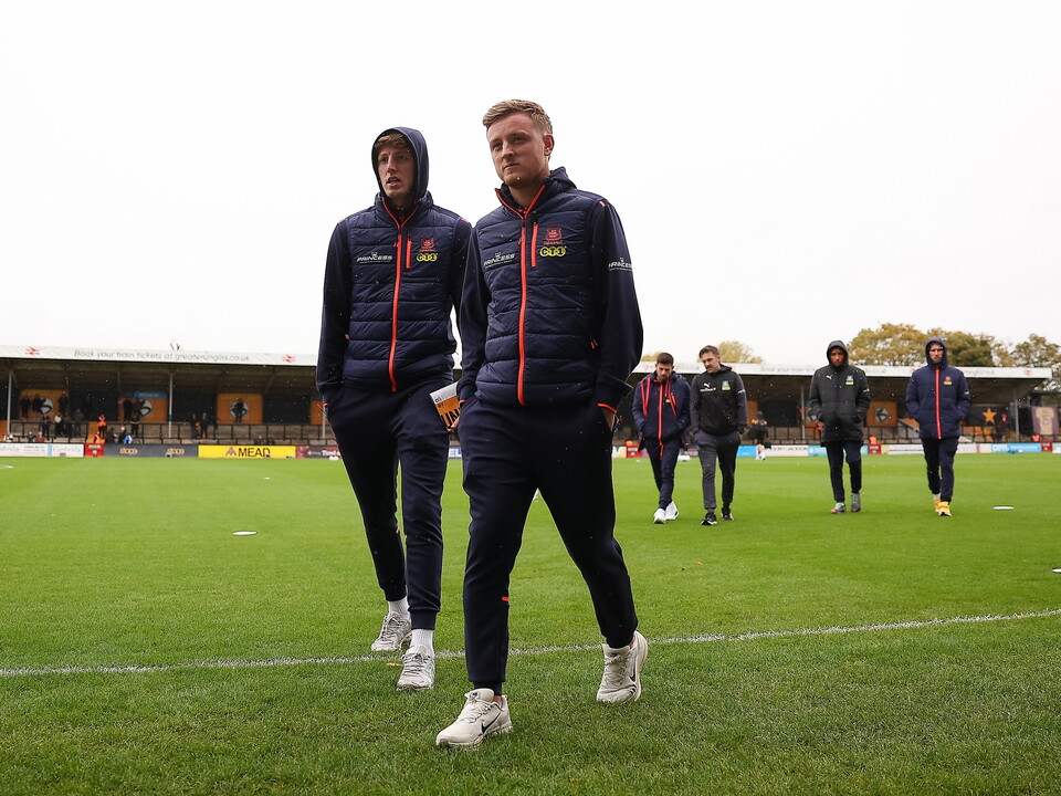 Two players in navy jackets and black pants walk across a football pitch.