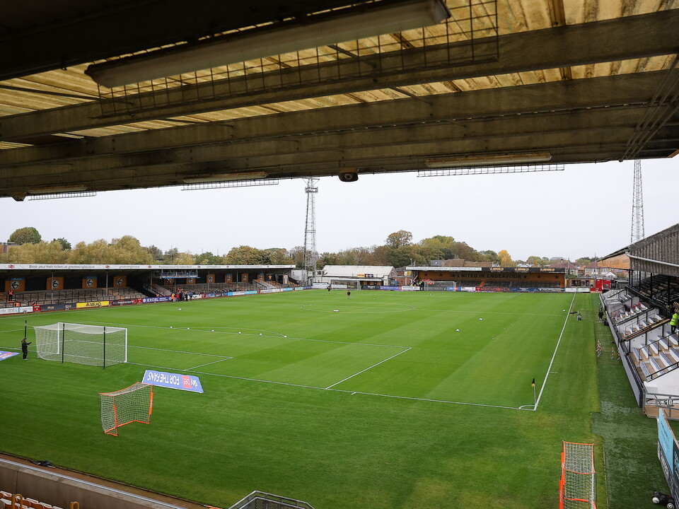An empty football stadium with a well-maintained green pitch and distant trees.