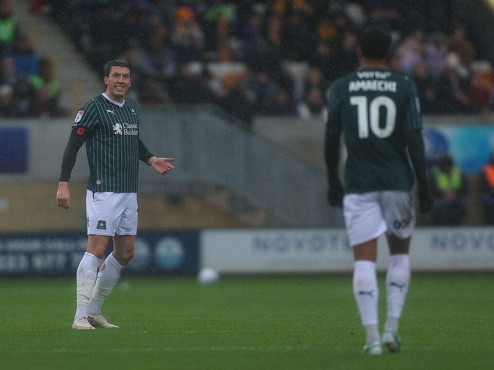 Two players in green shirts walk on a football field with a crowd watching.