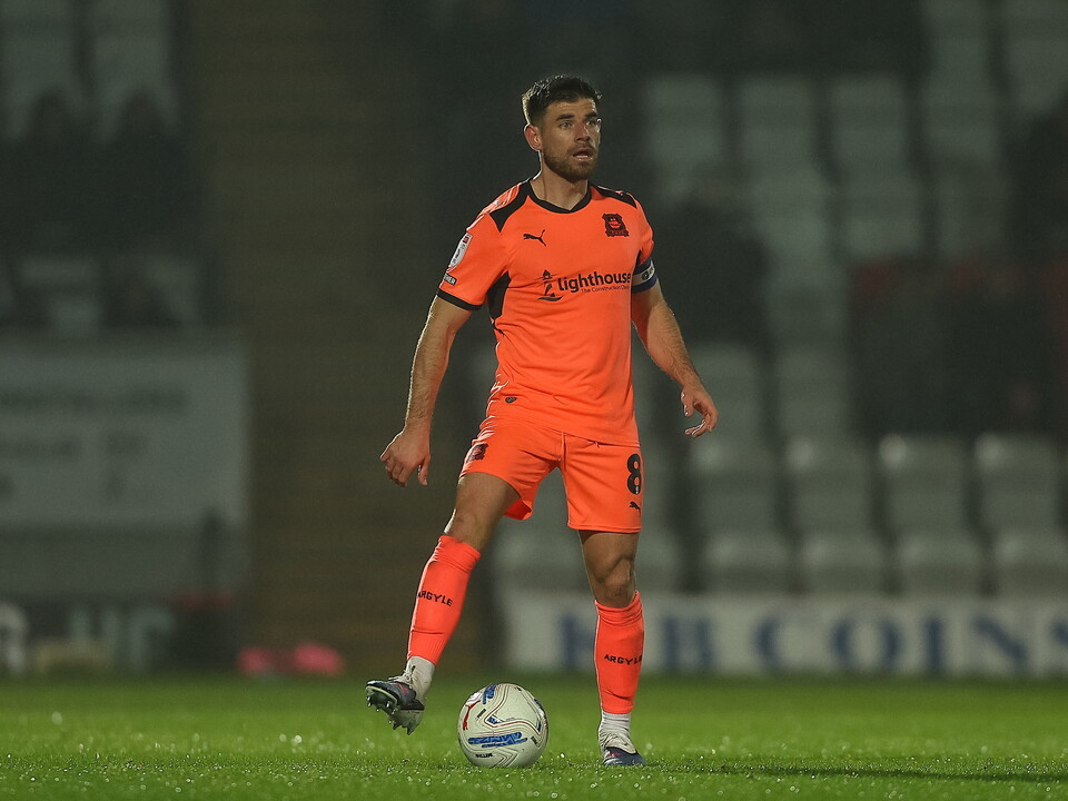 Footballer in orange kit kicks ball on green pitch, stadium background