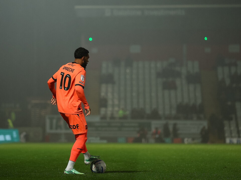 Football player number 10 in orange kit kicks ball on field, spectators in stands.