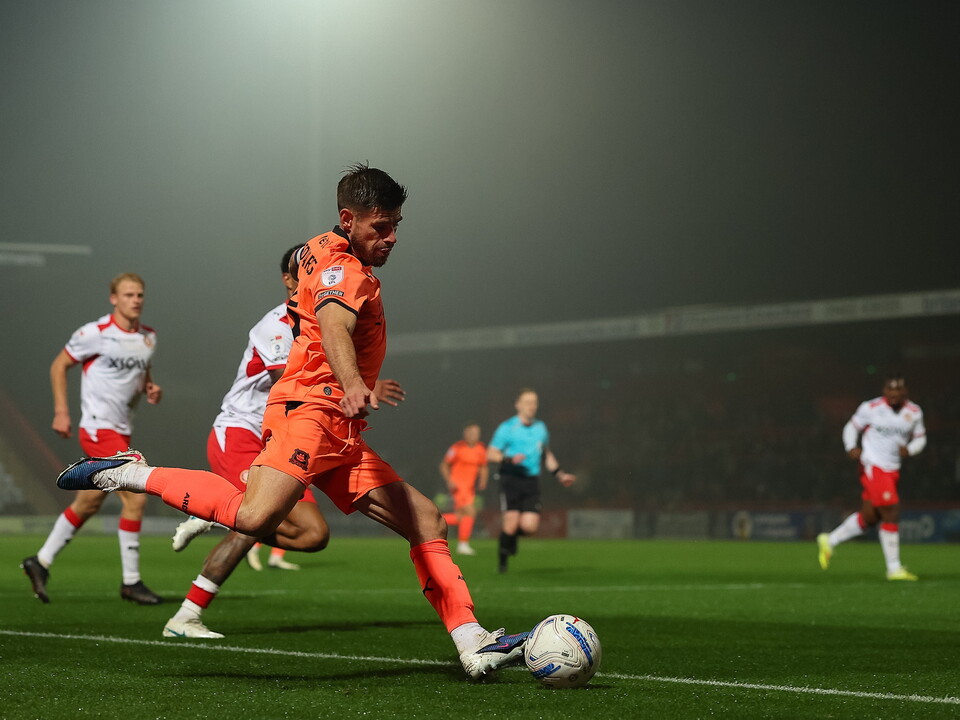 A footballer in orange kit slides to kick a ball on the pitch at night.