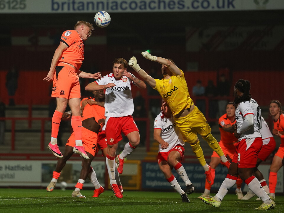 Players in orange, white, and yellow kits leap for the ball in a football match.