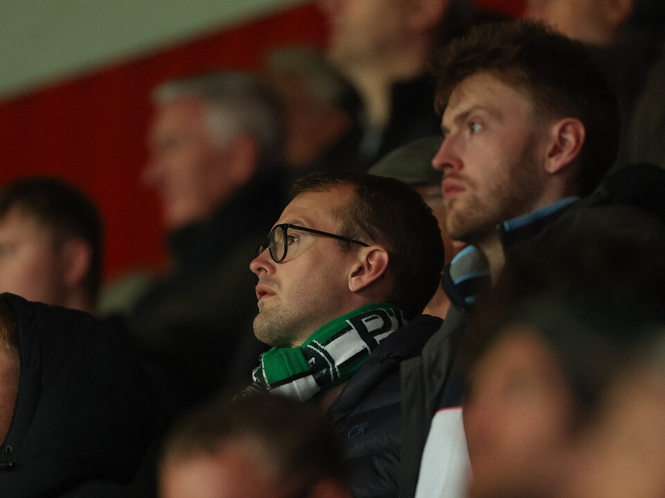 Fans wearing scarves, one with a green and white scarf, watch the game.