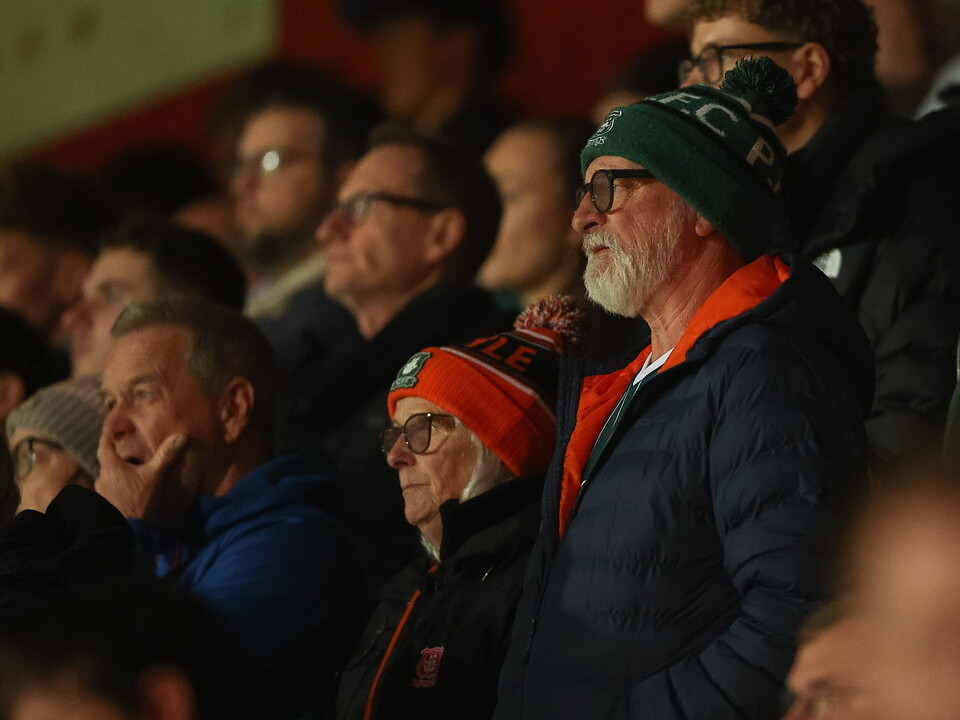 A crowd of football fans watching a match in a stadium, some wearing beanies and glasses.