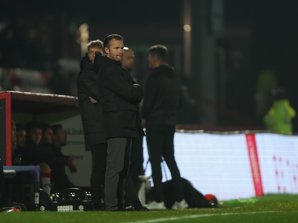 Four men in jackets, one with arms folded, stand on a football pitch under floodlights