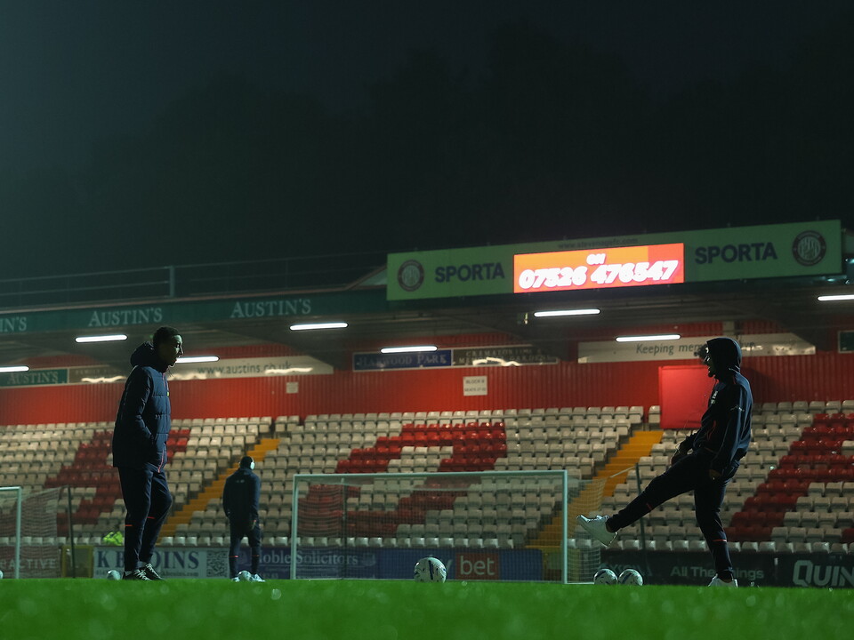 Three footballers in black training suits practice on a football pitch at night.