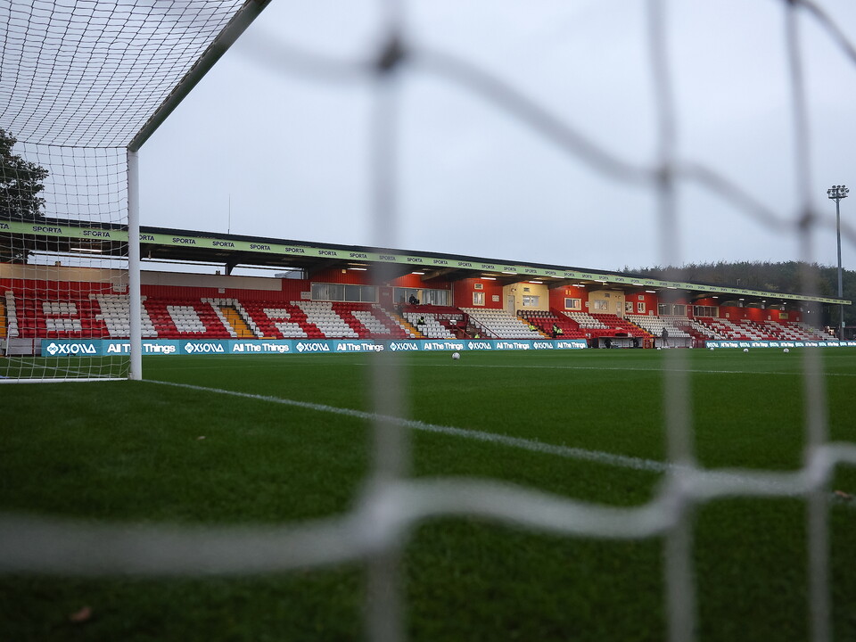 An empty football stadium with green pitch and red seats in the background.