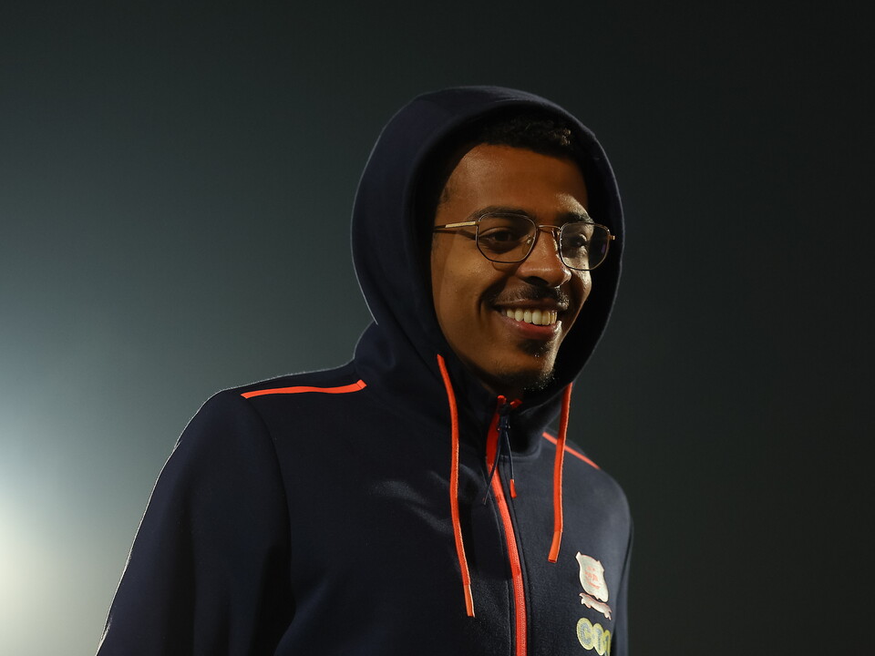 Man in navy blue hooded jacket with orange trims, wearing glasses, smiling.