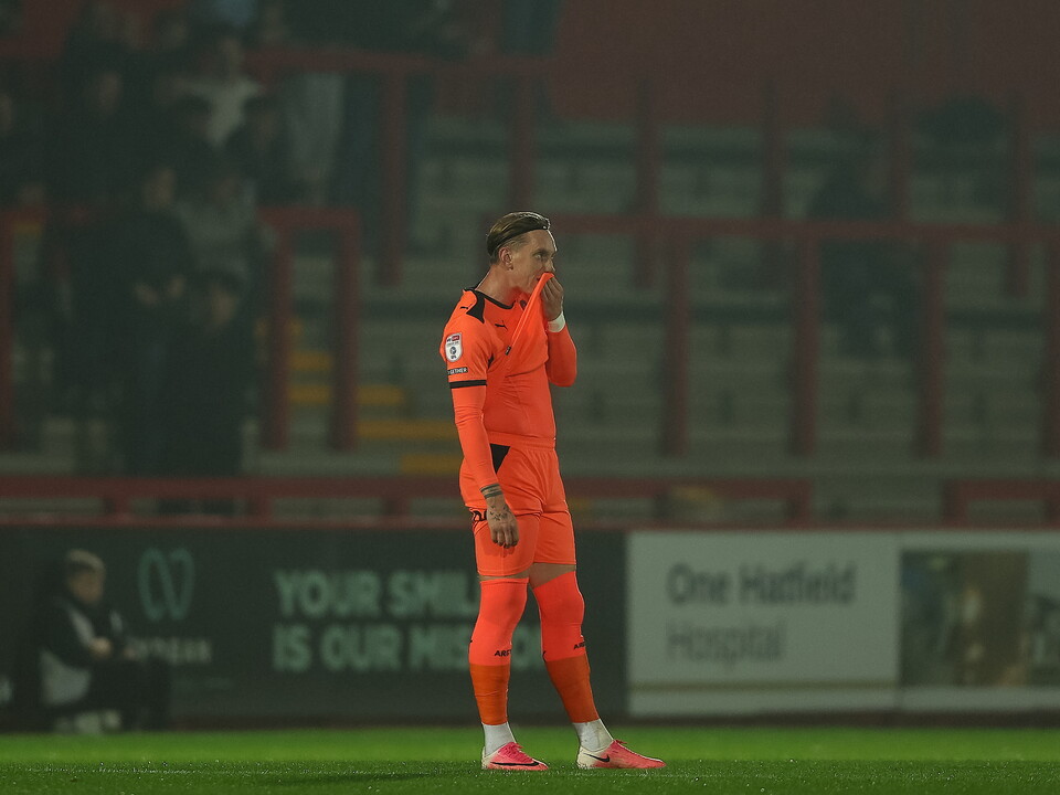 Football player in orange kit standing on pitch, hand on face, with blurred fans in stands.