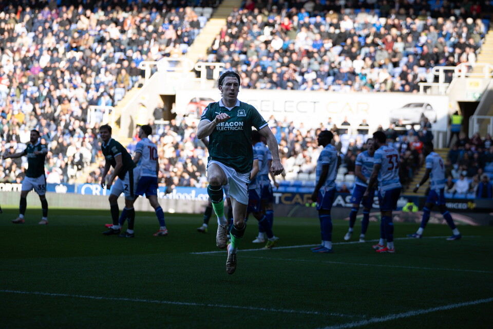 A football player in green running across a pitch with other players and a crowd behind.