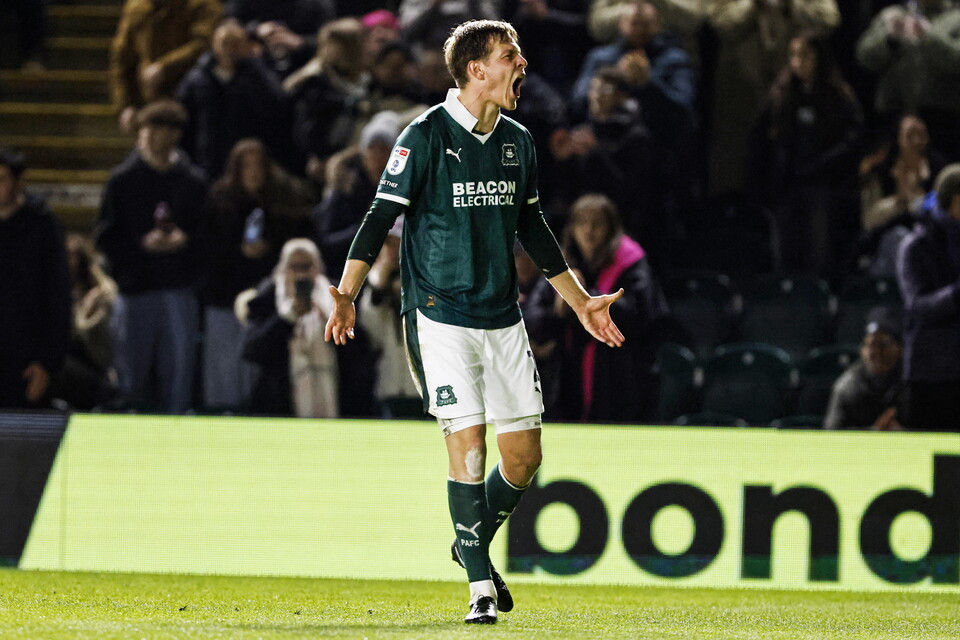 Soccer player in green jersey and white shorts walks on the field with arms raised.