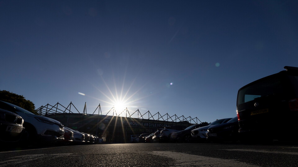 Bright sunlight over a car park with parked cars and stadium in the background.