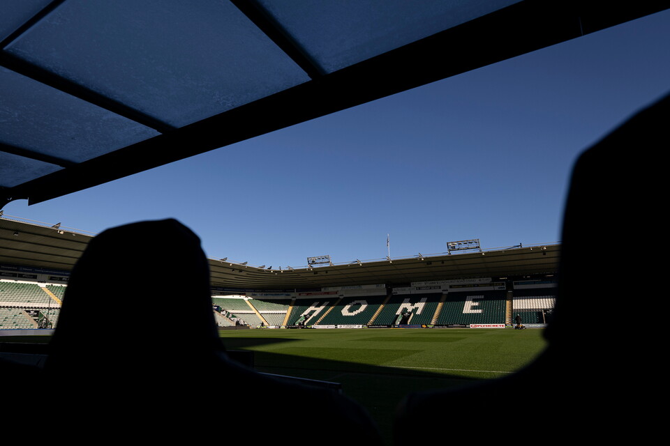 Two figures sit in a stadium with an empty pitch, the word "HOME" written in white on the opposite side.