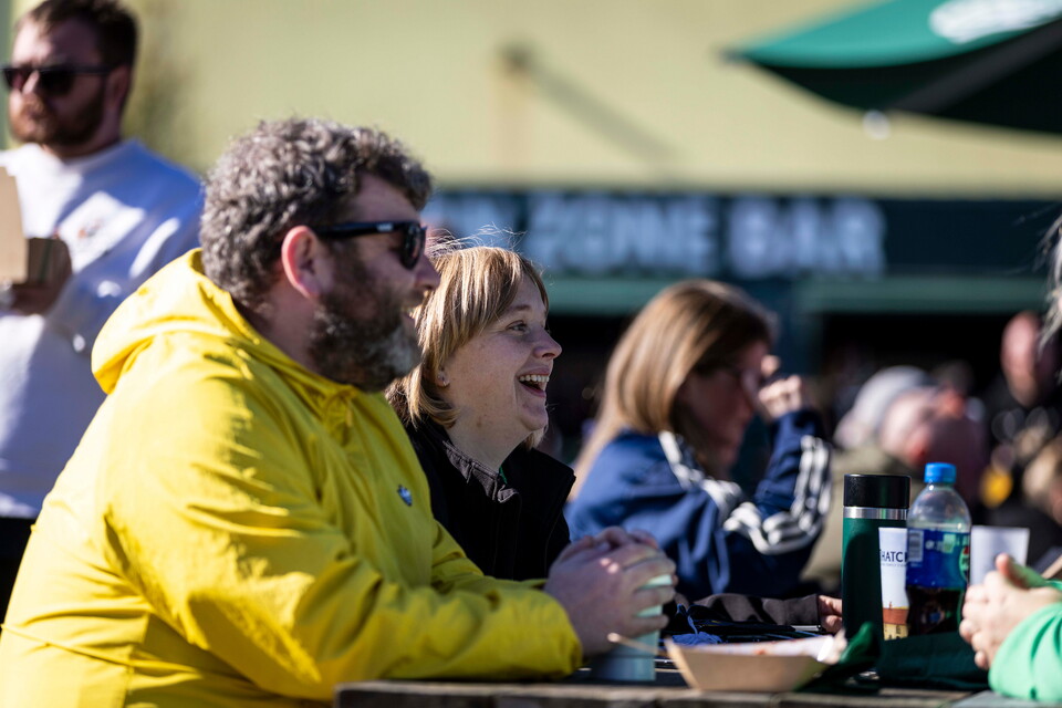 People in a bar, a man with a yellow hoodie laughs with two women, another woman has her hand on her chin.
