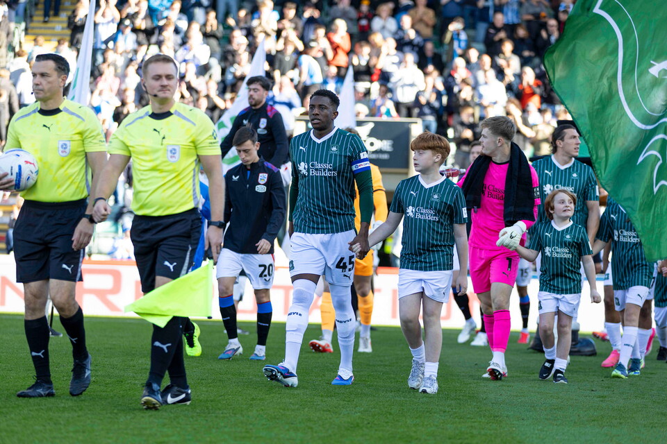 Football players walk off the pitch with children and supporters; some wear green shirts.