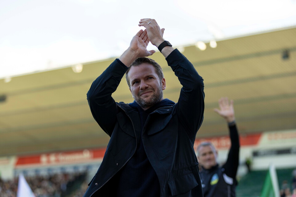 Man in black coat clapping hands while another man behind him raises a hand