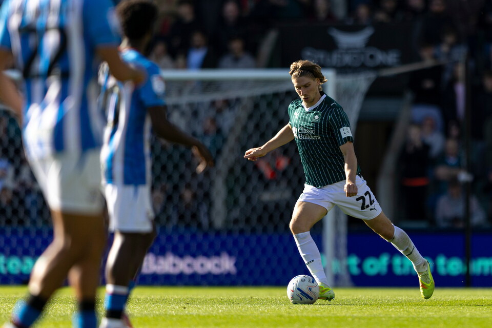 Footballer in green shirt kicks ball on pitch with player in blue and white shirt in background.