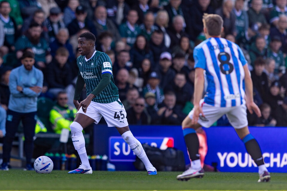 Footballer in green and white kit playing ball on field with a blue and white kit footballer behind