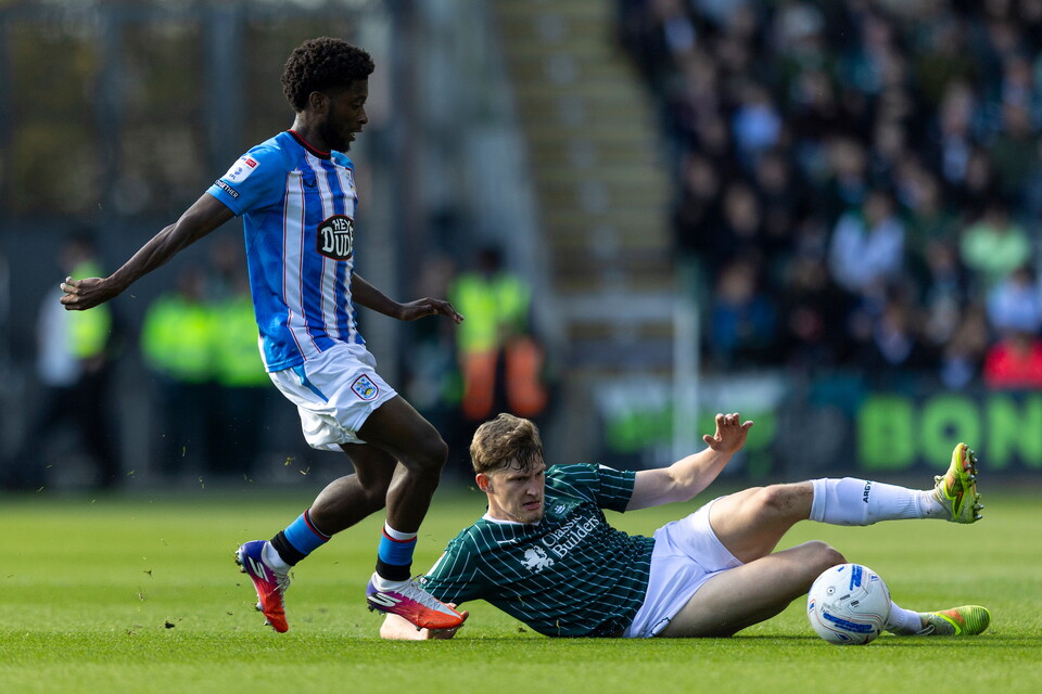 Footballer in blue slides past opponent in green; both players fall to ground.
