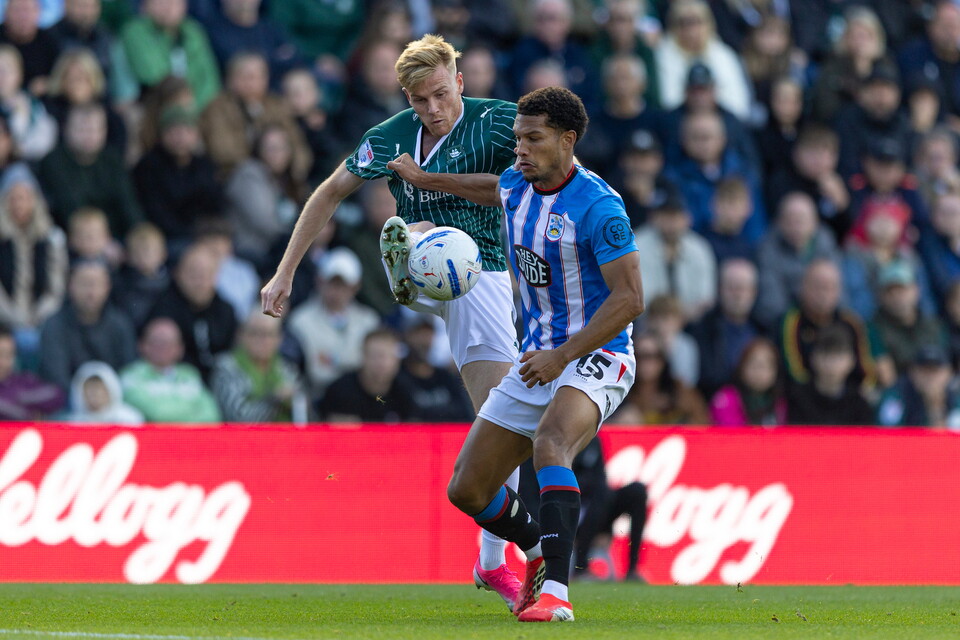 Two football players battle for the ball on a pitch in front of a crowd.