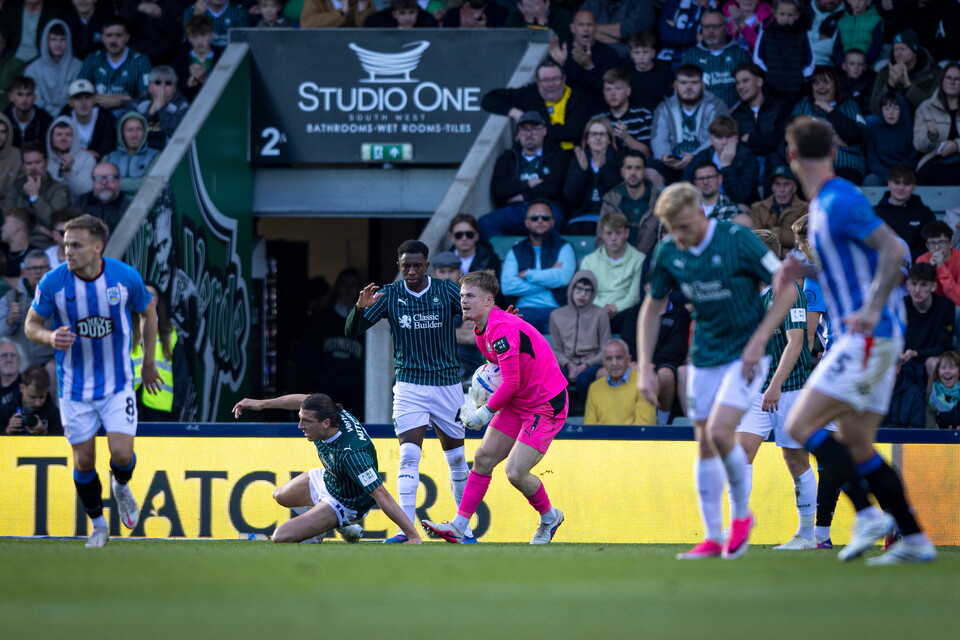 Soccer players compete on a pitch, one player sliding in front of a goalkeeper.