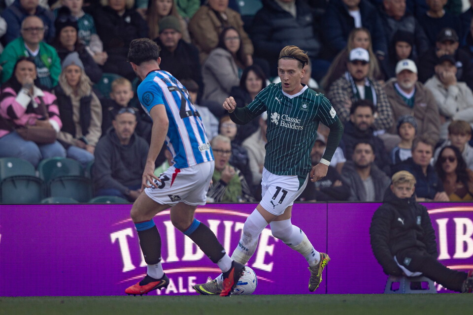 Two football players in blue and green jerseys run across a pitch in front of a crowd.