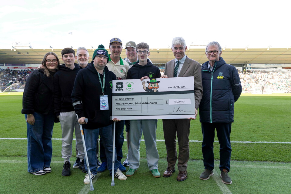 Nine people stand on a football pitch holding a cheque for 21,100 British Pounds.