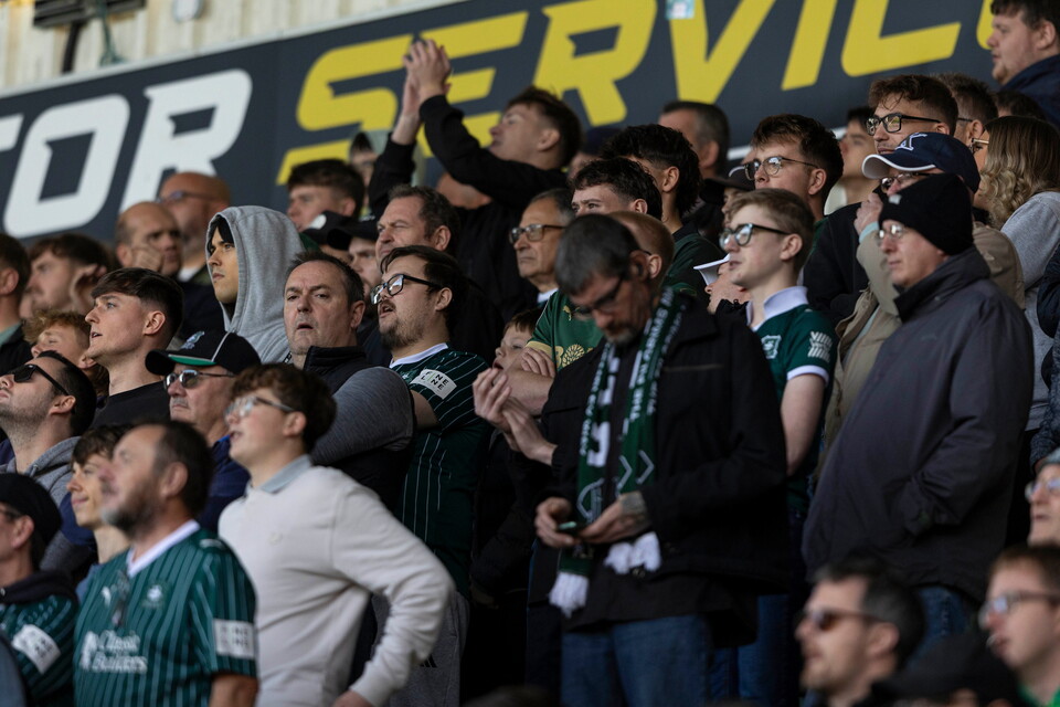 A crowd of people in a stadium, some wearing green and black shirts and others in casual clothing, cheering during a football match.