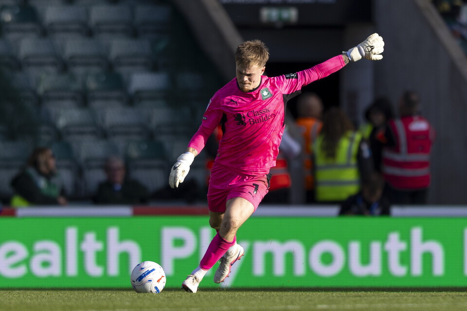Player in pink kit kicks soccer ball on field, spectators behind ad board.