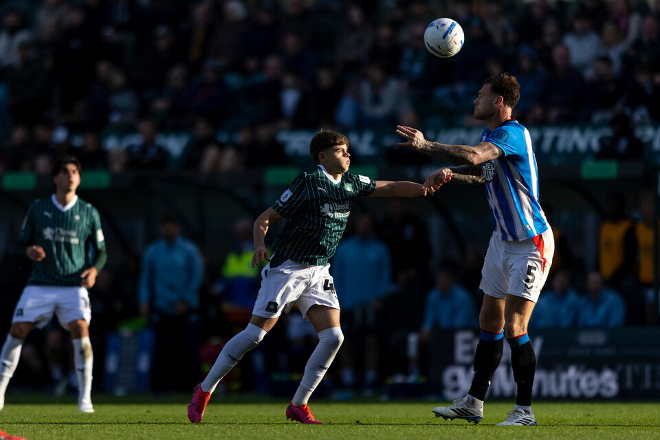 Two football players in action on a pitch, one in green and white, the other in blue and white.