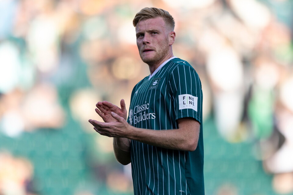 Footballer in green striped shirt claps hands, standing on green pitch, crowd in background.