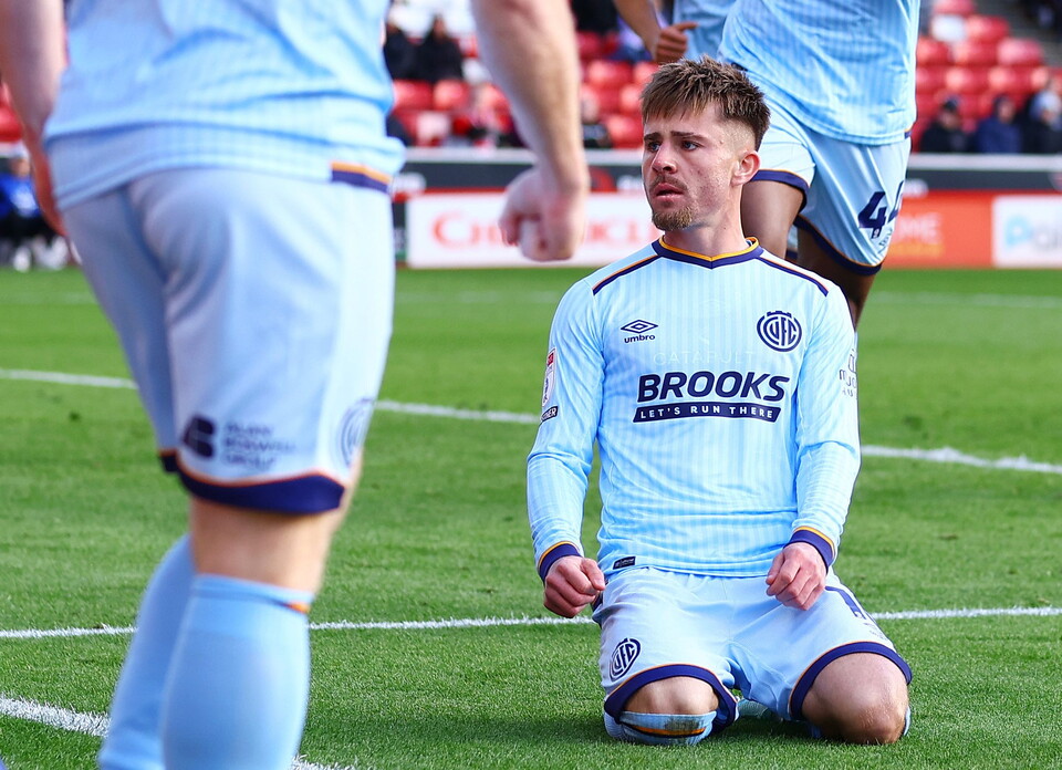 Footballer wearing light blue shirt with Brooks logo kneels on field, looking disappointed