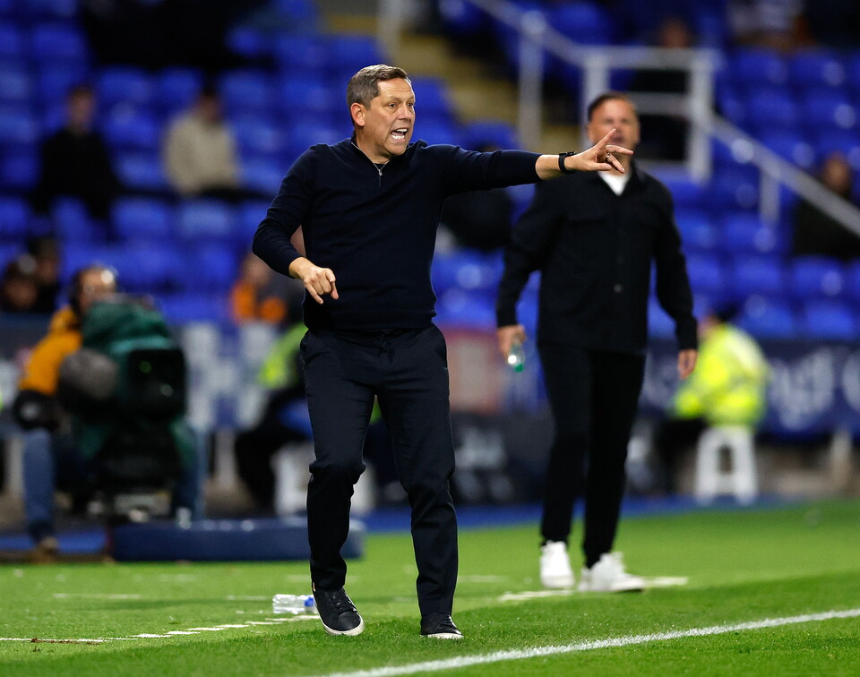 A coach gestures on a football pitch, with another coach and an assistant standing behind him.