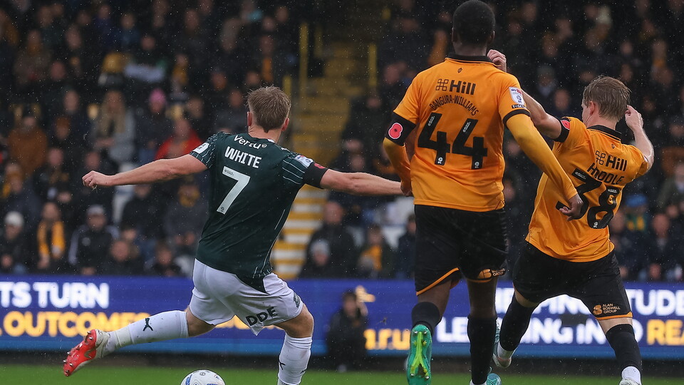 Three football players in action, one in green shirt with number 7, two in orange shirts.