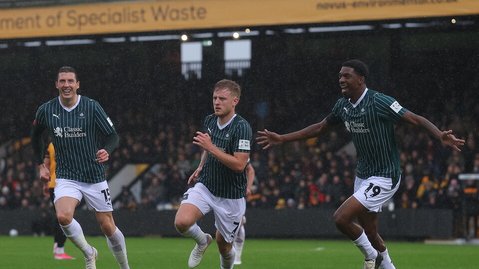 Three players in green shirts and white shorts celebrate a goal on a football pitch.