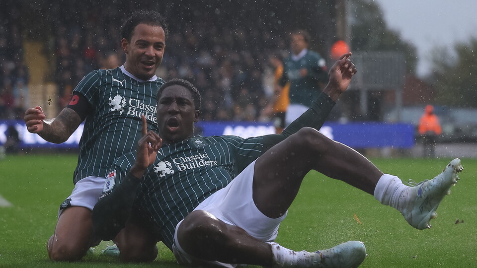Two players in green and white kits, one celebrating a goal with arms raised, the other sliding on a wet pitch.