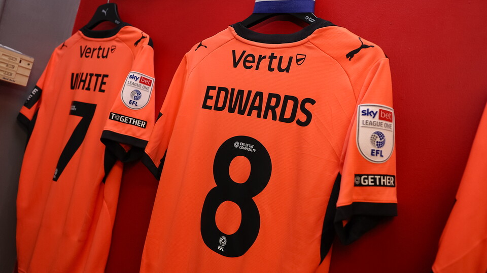 Three orange football shirts with names and numbers hanging on a red wall.