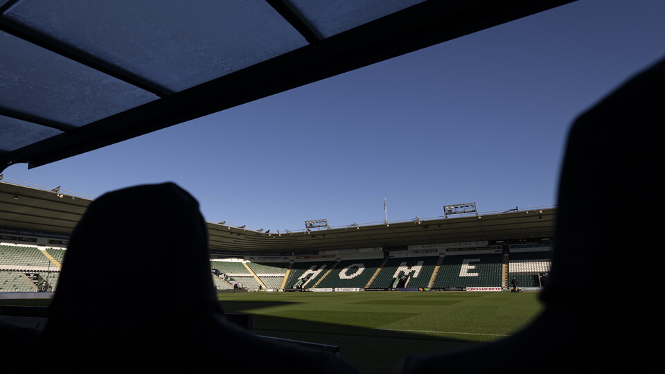 Two silhouettes of people looking out at an empty football stadium with a green pitch.