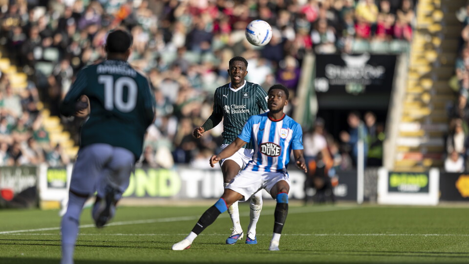 Two football players in green and blue challenge for a ball in midair on a pitch.