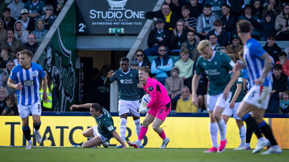 Players in green, pink, and blue uniforms compete in a football match; spectators watch from the stands.