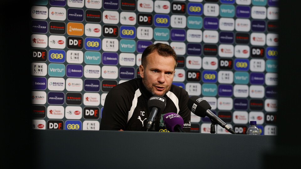 A man in a black and white shirt is giving a press conference in front of a microphone.
