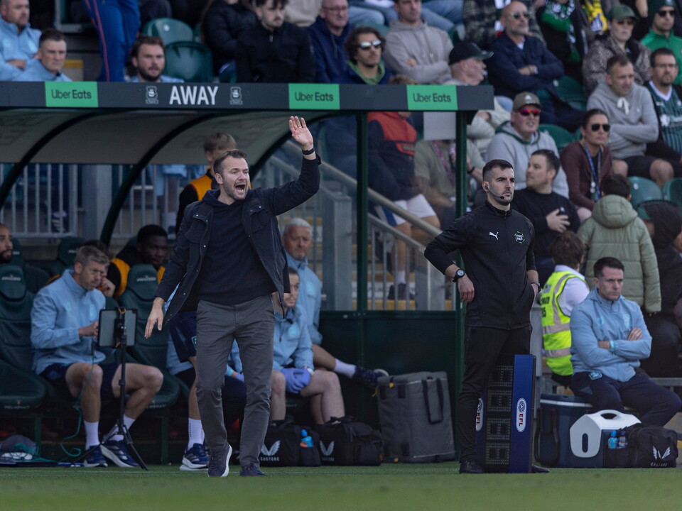 Football coach gesturing to players in a blue and white striped shirt on a pitch.